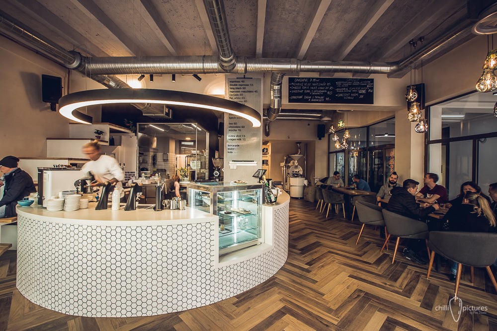 The curved white hexagon-tiled counter and open roastery-style interior at Lékárna by City Roasters, with a barista in motion, a herringbone wood floor, and guests seated at communal tables